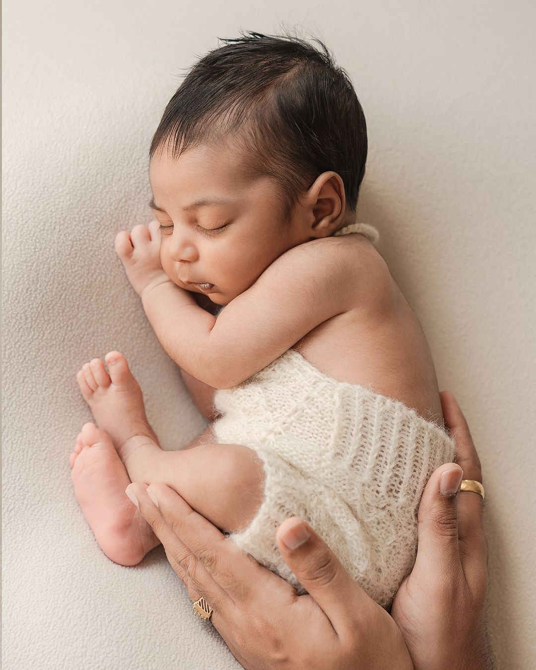 Sleeping newborn in cream knit shorts curled up while parent gently supports tiny feet.