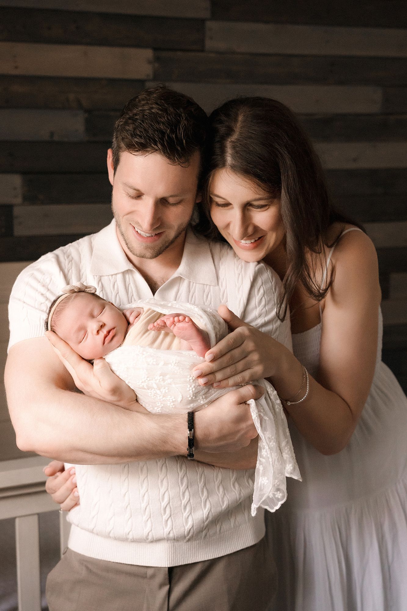 Smiling couple in white outfits cradling their sleeping newborn baby wrapped in lace.