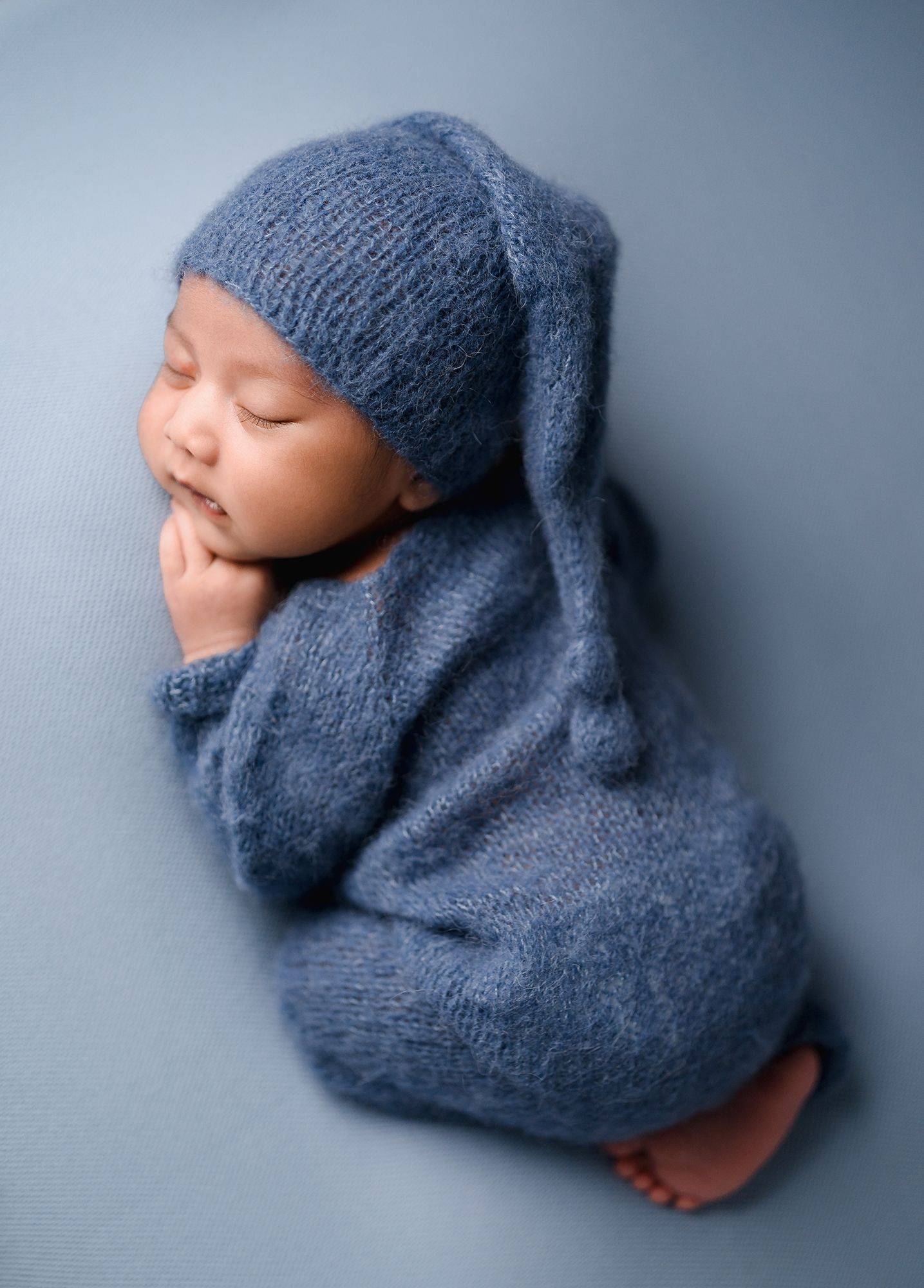 Sleeping newborn baby in a blue knit romper and long-tailed hat curled up on a soft blue backdrop in a newborn photography portrait.