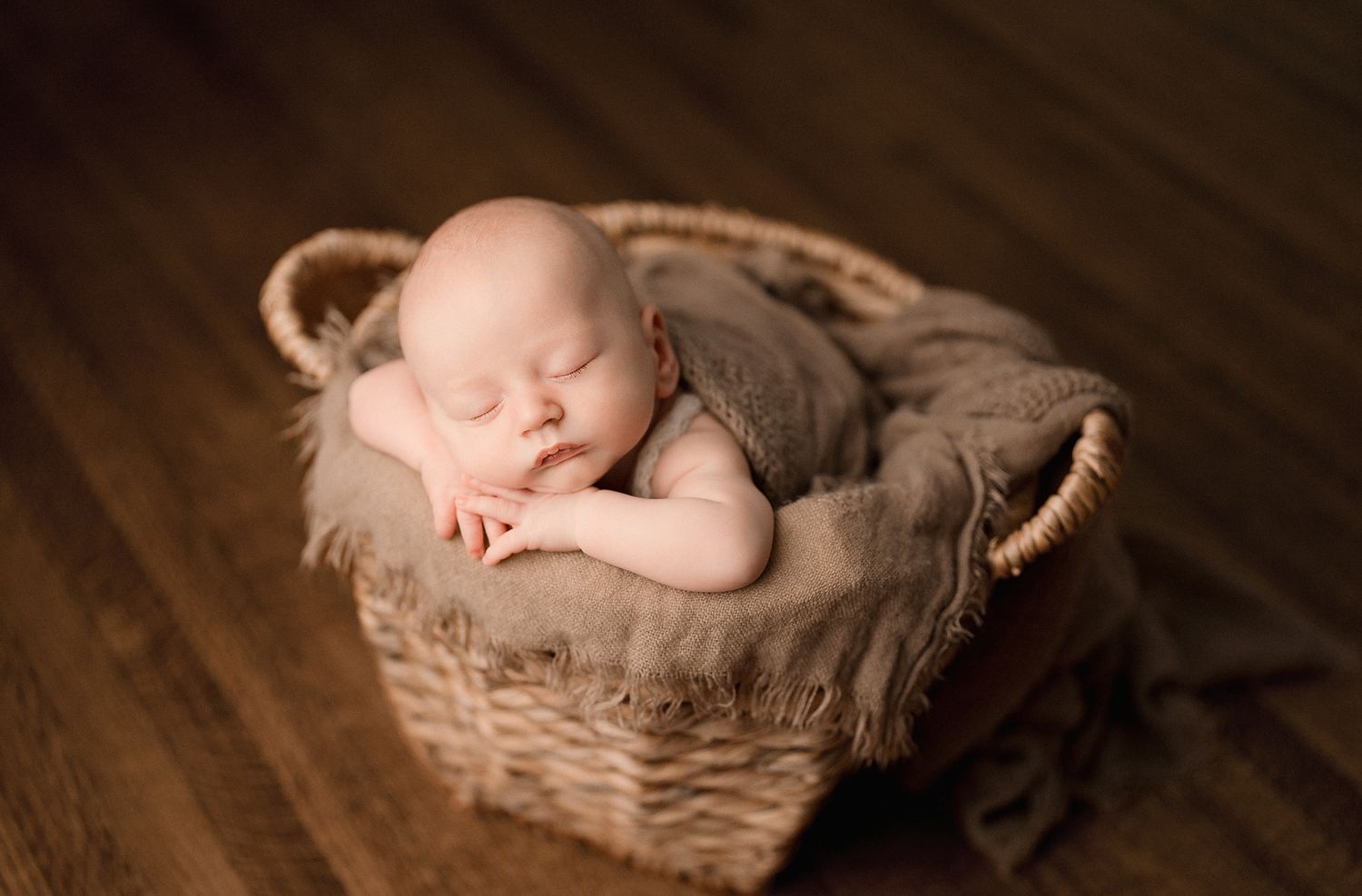 A peaceful newborn baby sleeps curled up in a wicker basket with soft brown blanket.