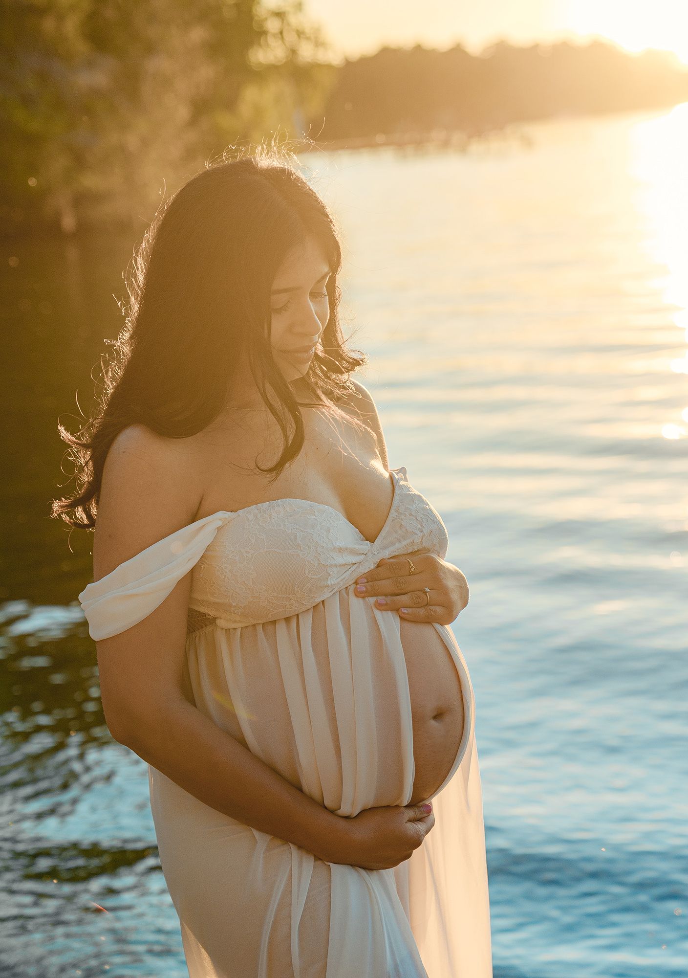 Pregnant woman in white off-shoulder gown cradling her baby bump at golden sunset by the water.