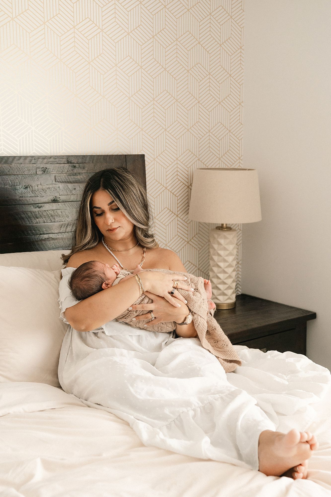New mother in a white dress sits on a bed cradling her sleeping newborn baby in a bright bedroom with geometric wallpaper.