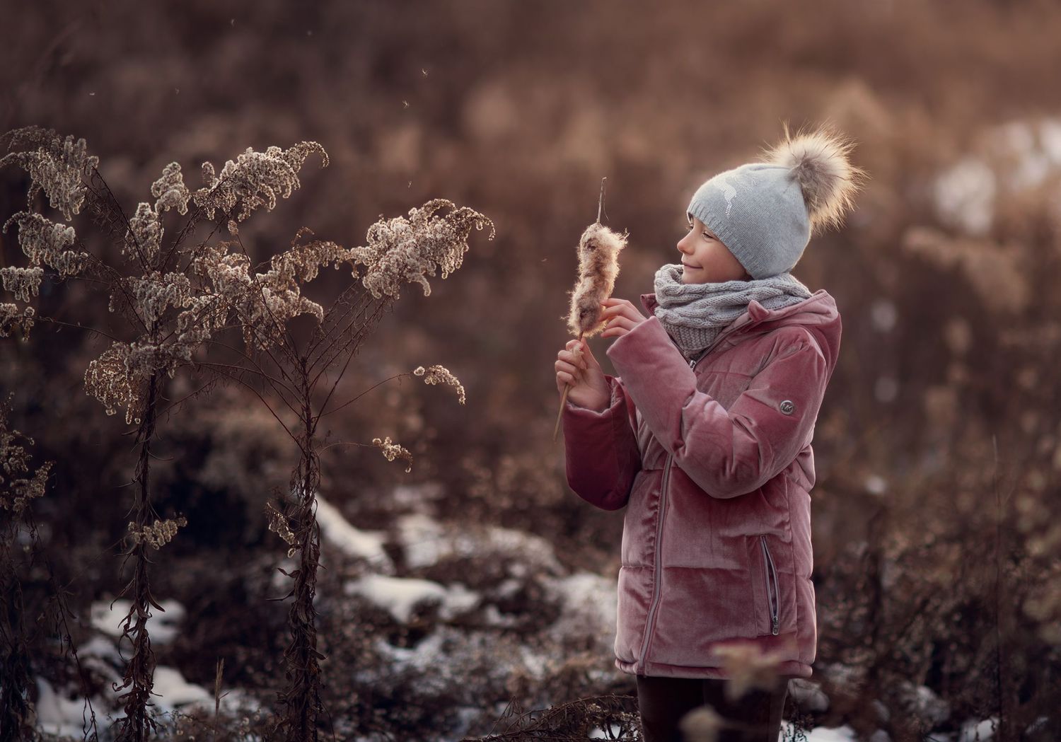 A young girl in a pink winter coat and gray hat explores frosty plants in a snowy field.