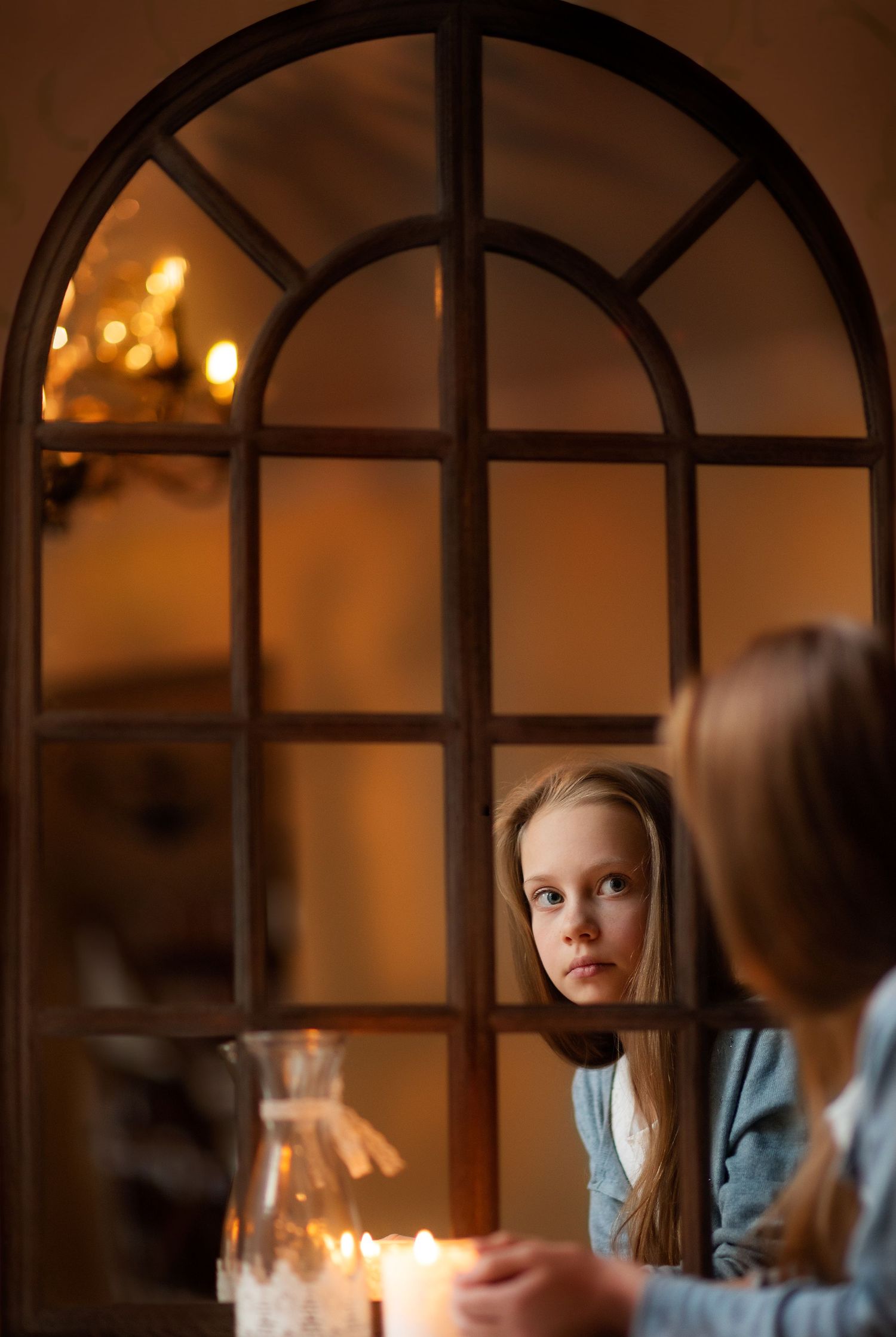 A young blonde girl peers through an arched window frame with candlelight glowing in a warmly lit room.
