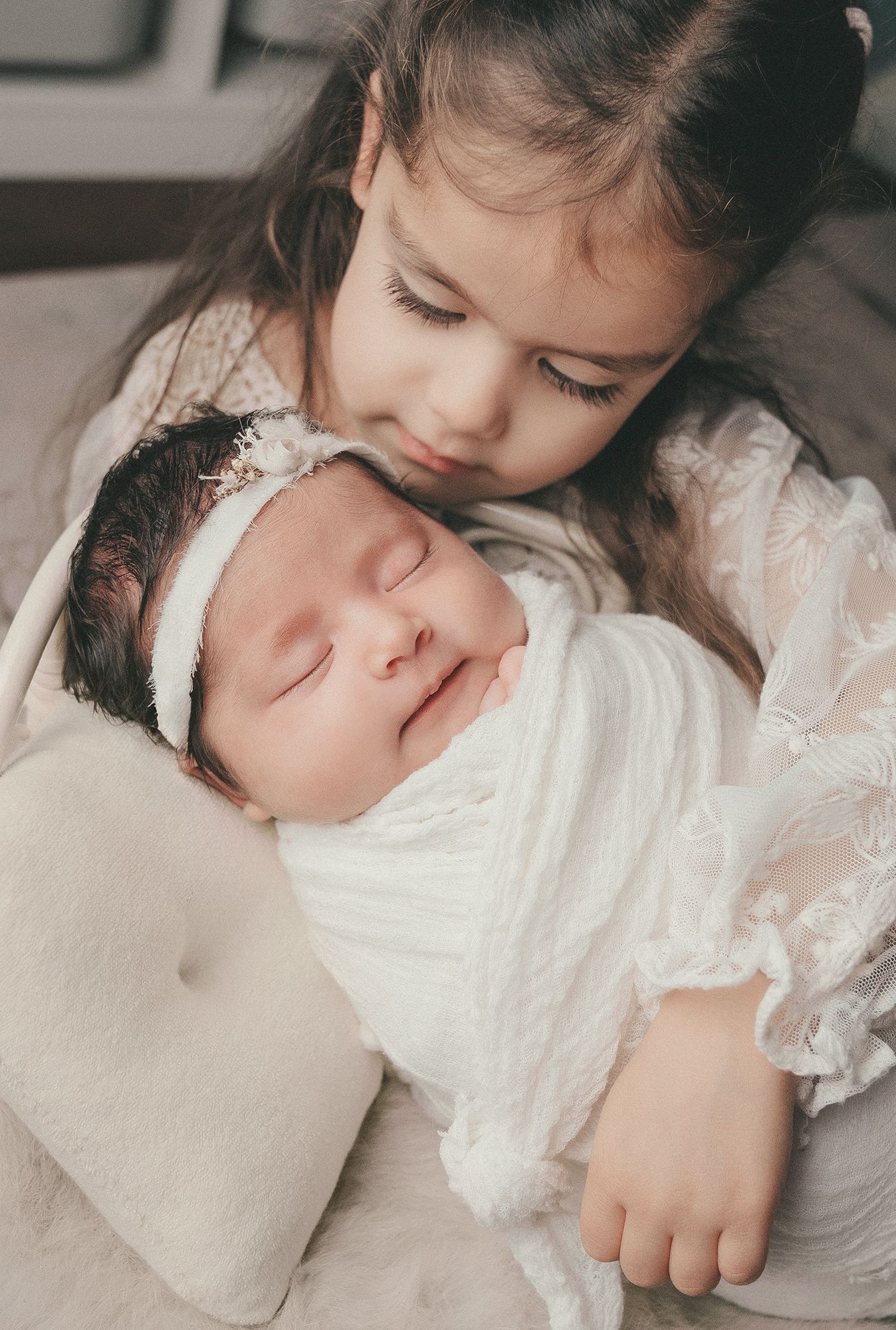 A young girl lovingly kisses her sleeping newborn sibling wrapped in a white swaddle blanket.