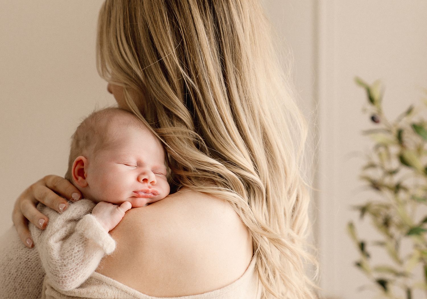 Mother with long blonde hair holding a sleeping newborn baby against her shoulder in a soft, neutral-toned room.
