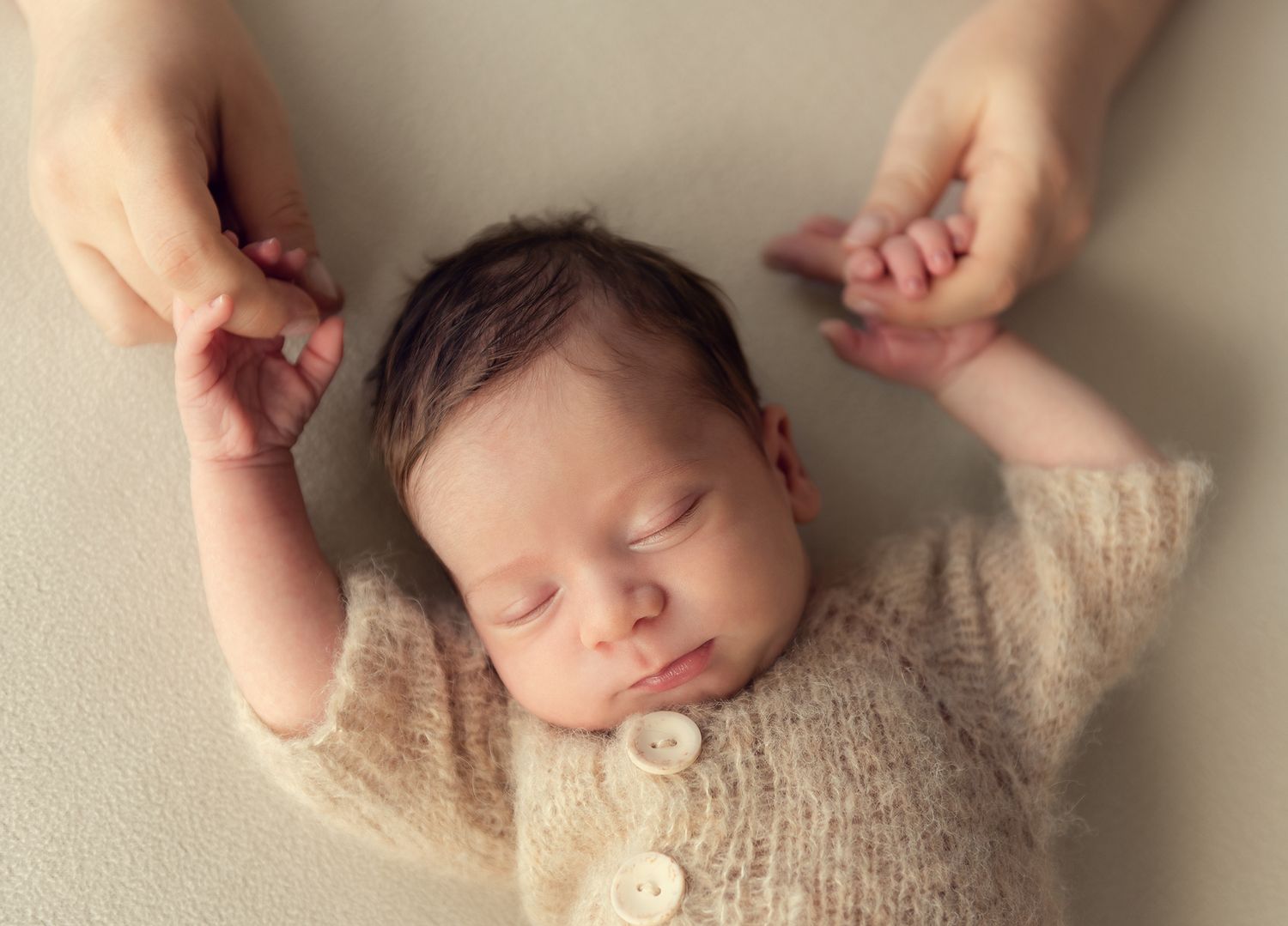 Sleeping newborn in a cream knit cardigan gently held by parents hands from above.