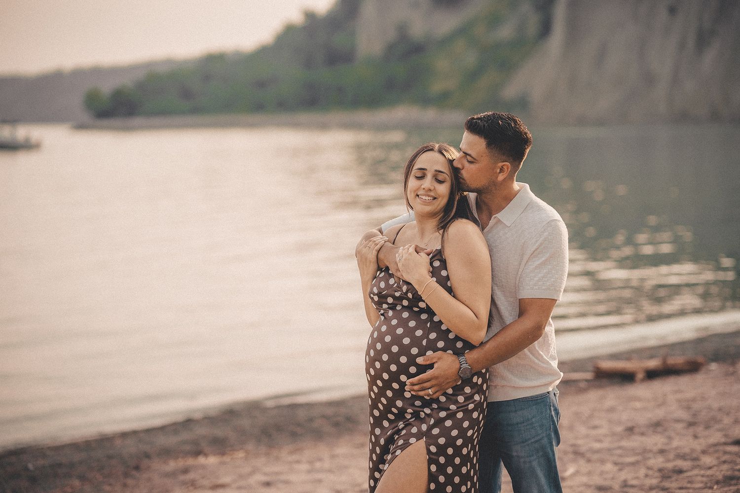 Couple embracing on rocky beach at sunset, woman in polka dot maternity dress.