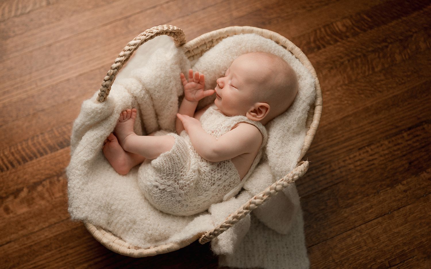 Newborn baby sleeping peacefully in a wicker basket lined with soft cream blanket on a wooden floor.