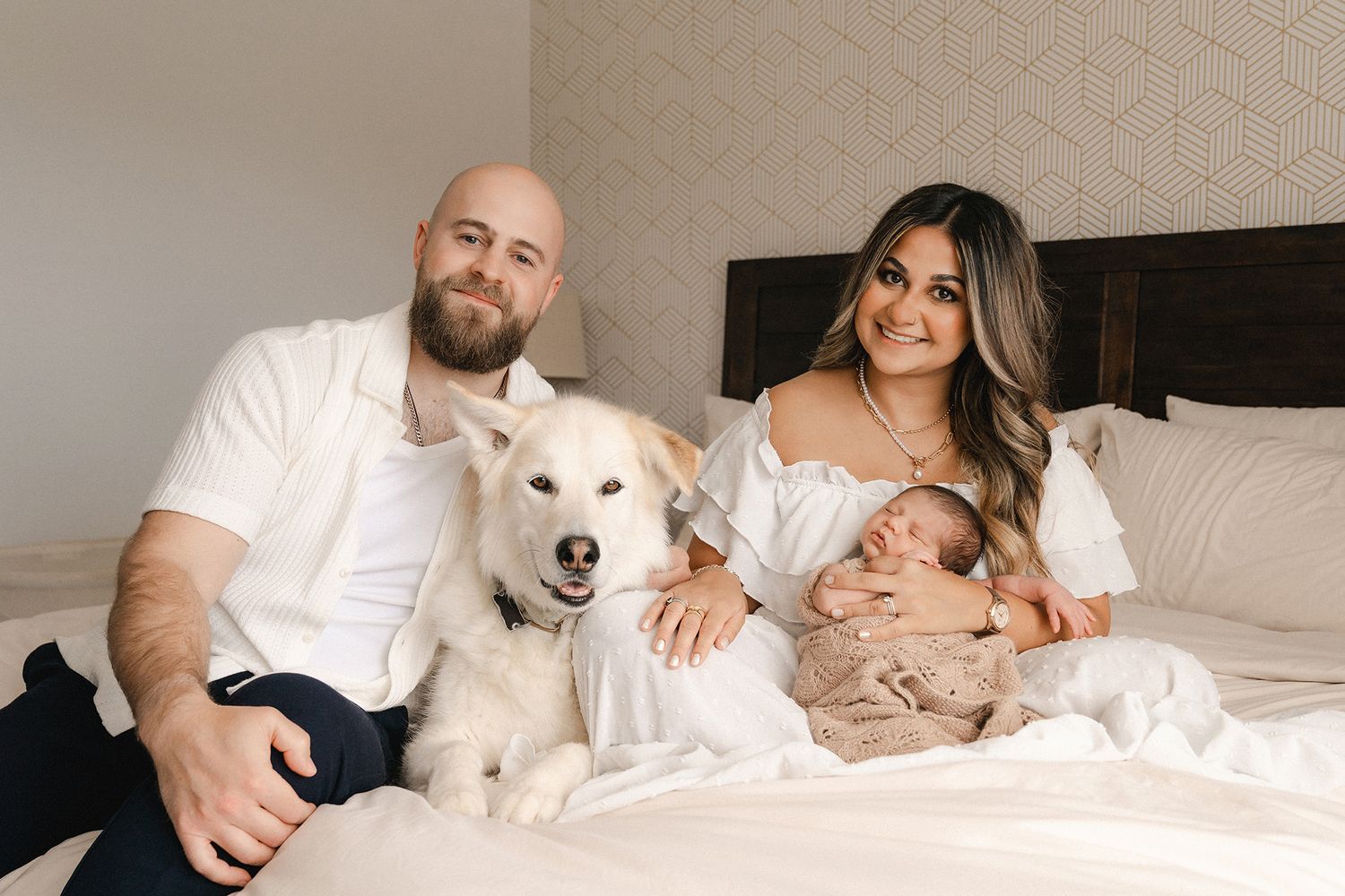 A smiling man and woman sit on a bed with a fluffy white dog while the woman cradles a sleeping newborn baby in a cozy bedroom.