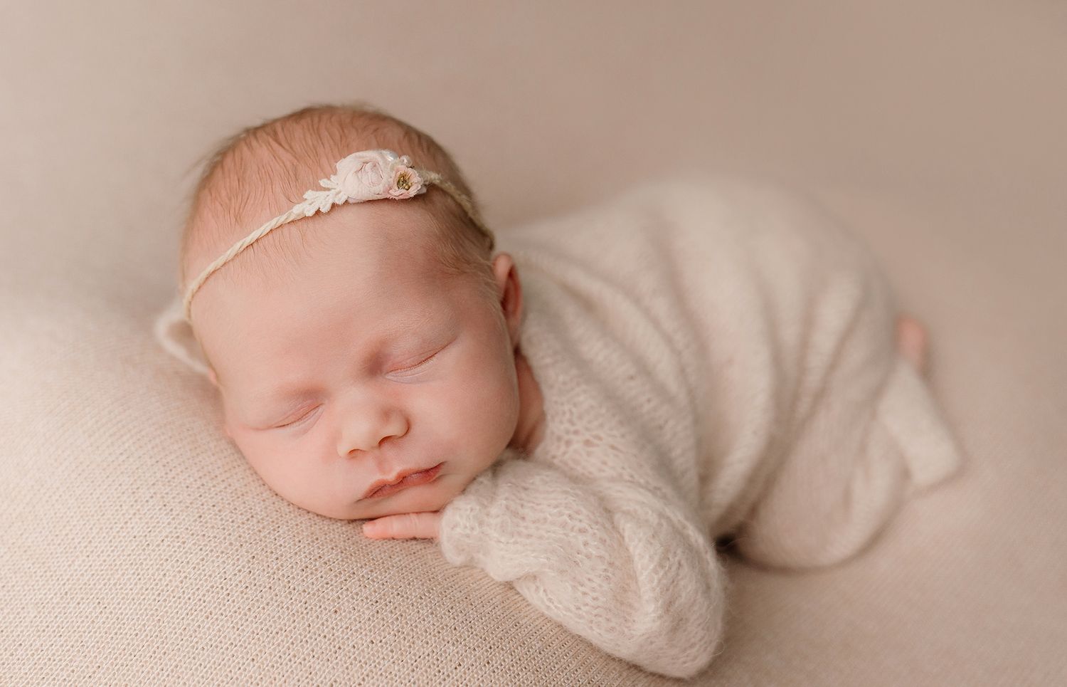 Sleeping newborn baby in a cream knit outfit and floral headband resting peacefully on a soft neutral blanket.