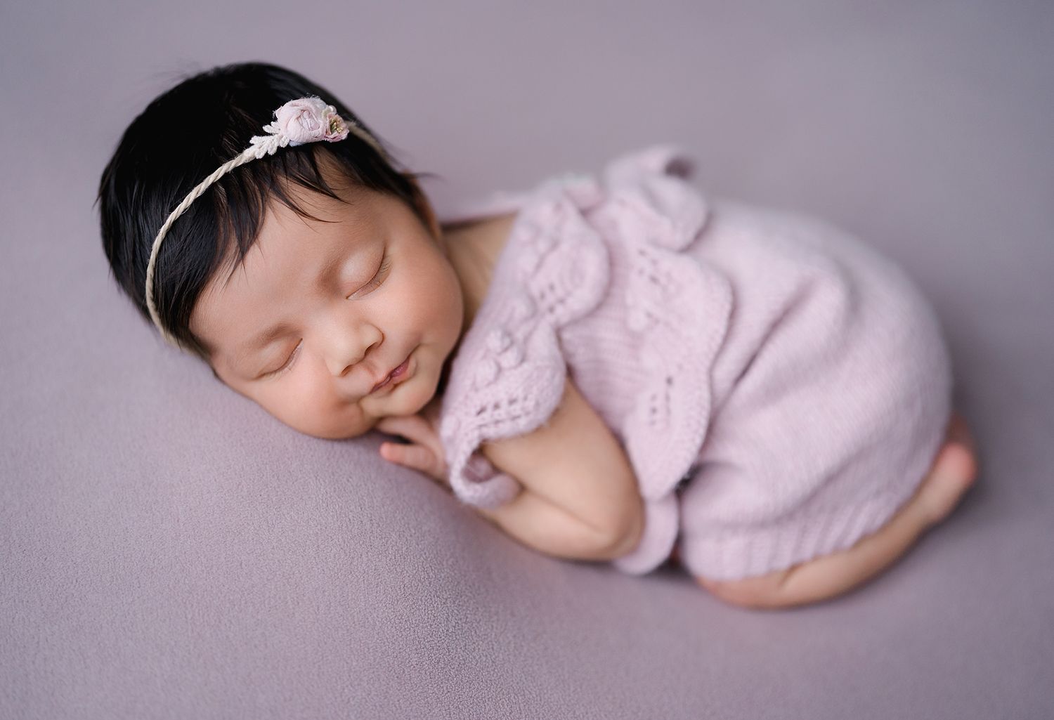 A serene newborn baby girl sleeps peacefully in a lavender knit outfit on a matching backdrop.