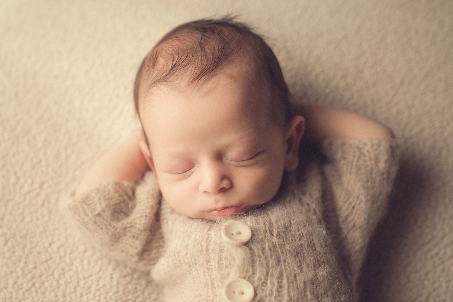 Sleeping newborn baby in a beige knit onesie with buttons lying peacefully on a soft cream blanket.