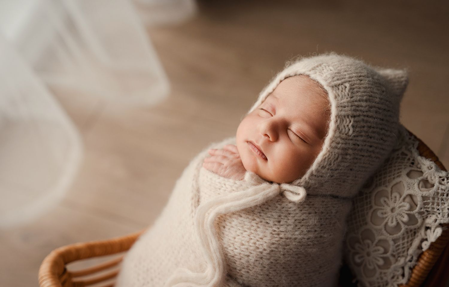 Sleeping newborn baby swaddled in cream knit wrap with bonnet resting in a wicker basket.
