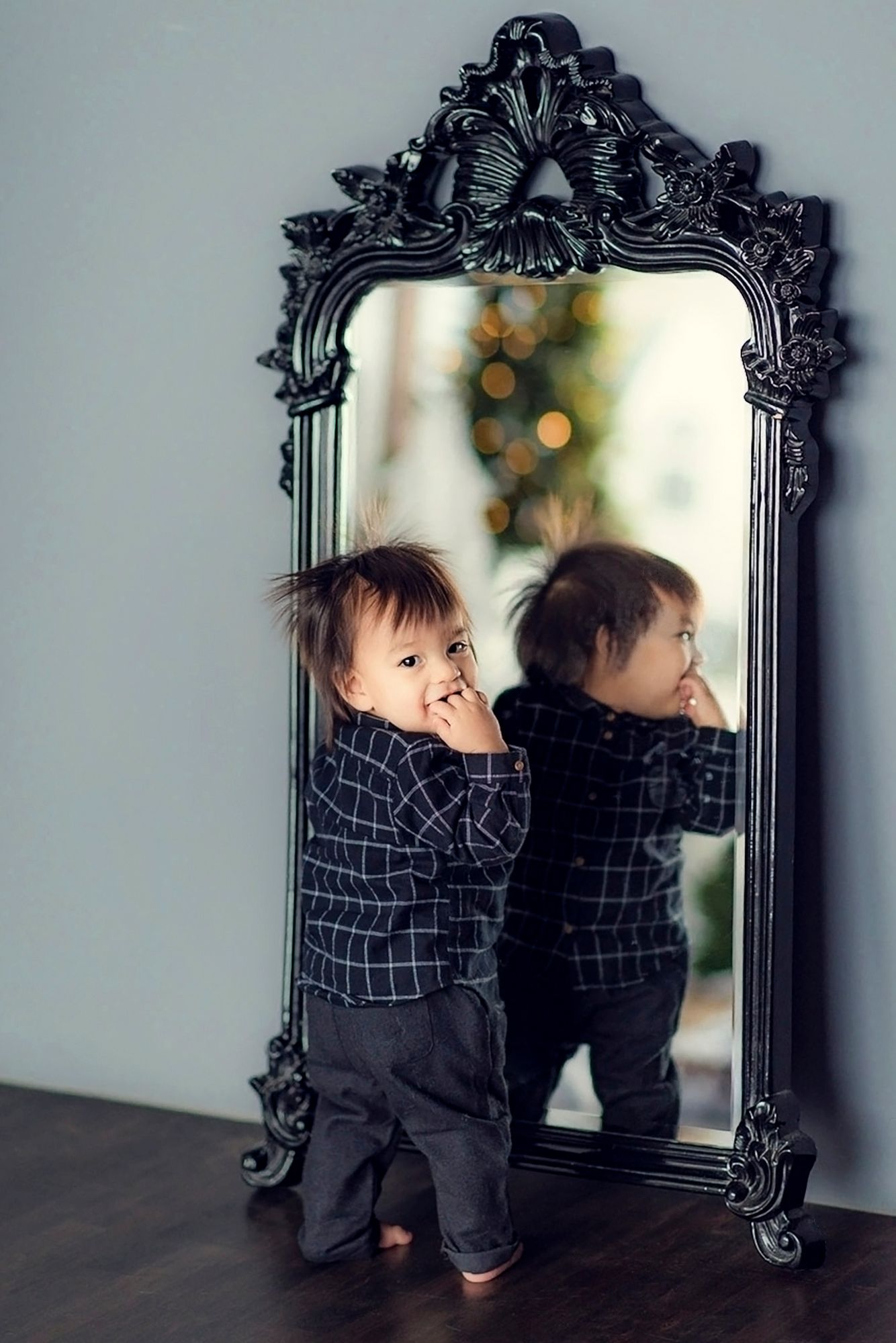 An adorable toddler in a checkered shirt smiles at his reflection in a decorative black mirror.