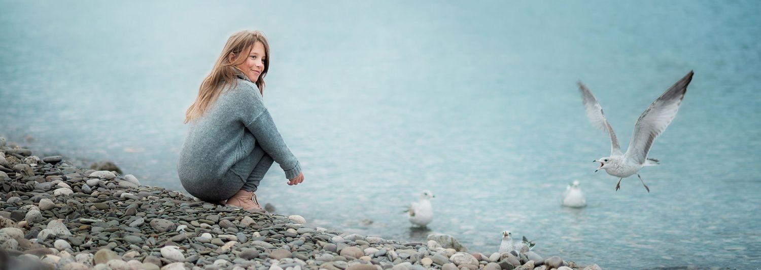 Serene woman sitting on pebble beach in blue outfit observing seagulls gliding over tranquil water.