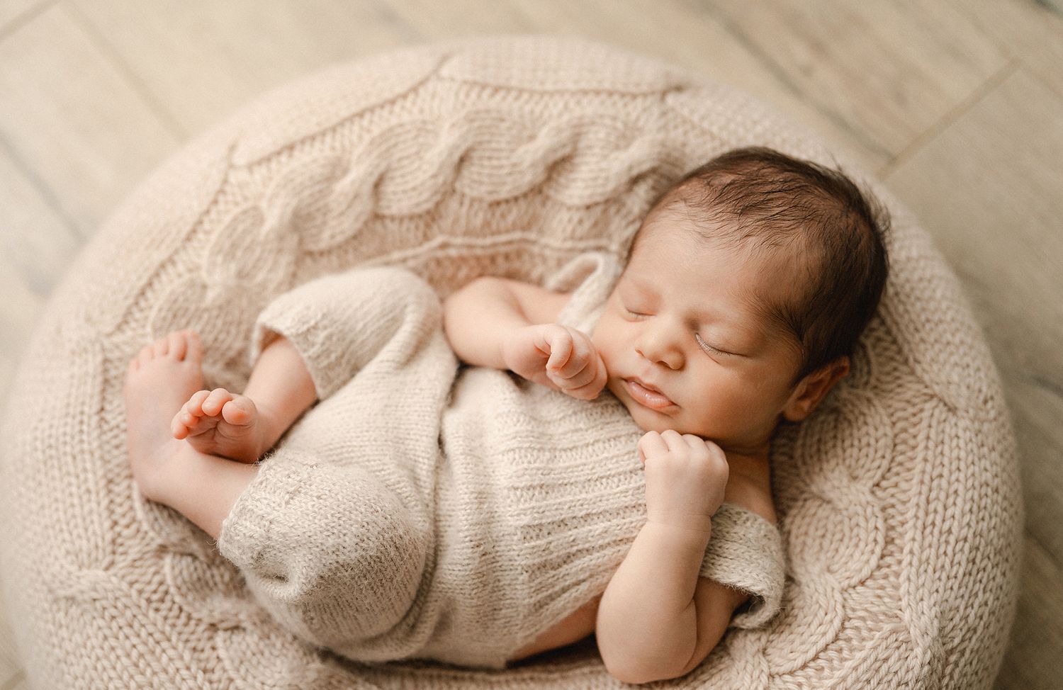 Sleeping newborn baby in a cream knit romper curled up on a cozy cable-knit posing pouf during a soft, neutral newborn photoshoot.