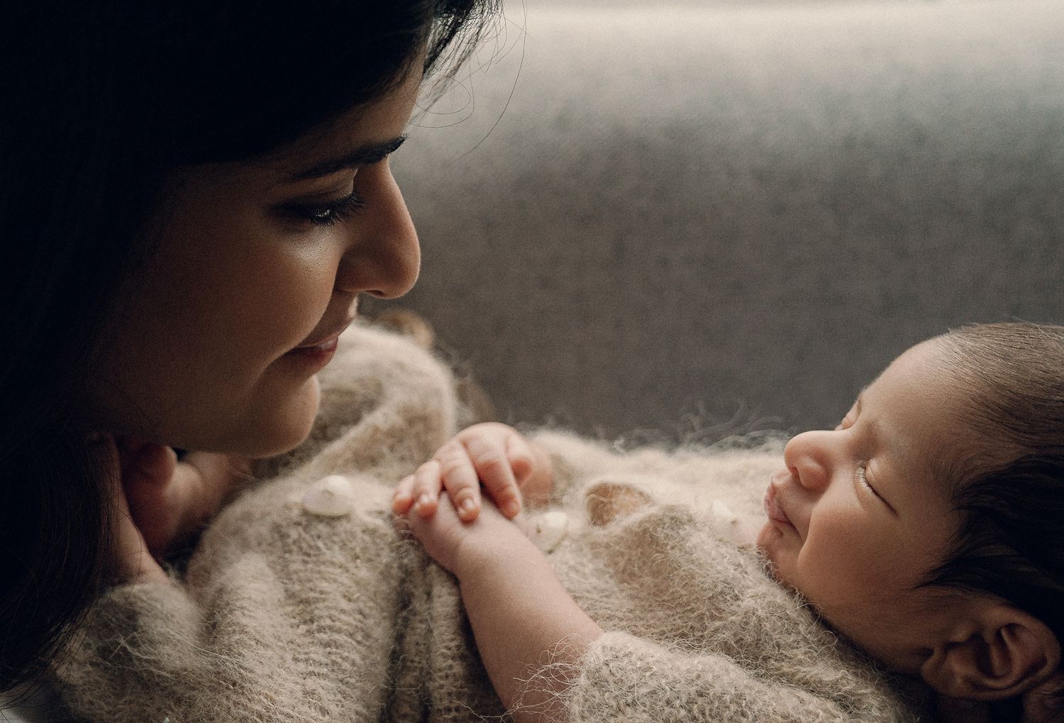 A loving mother gazes at her sleeping newborn baby wrapped in a soft beige blanket.