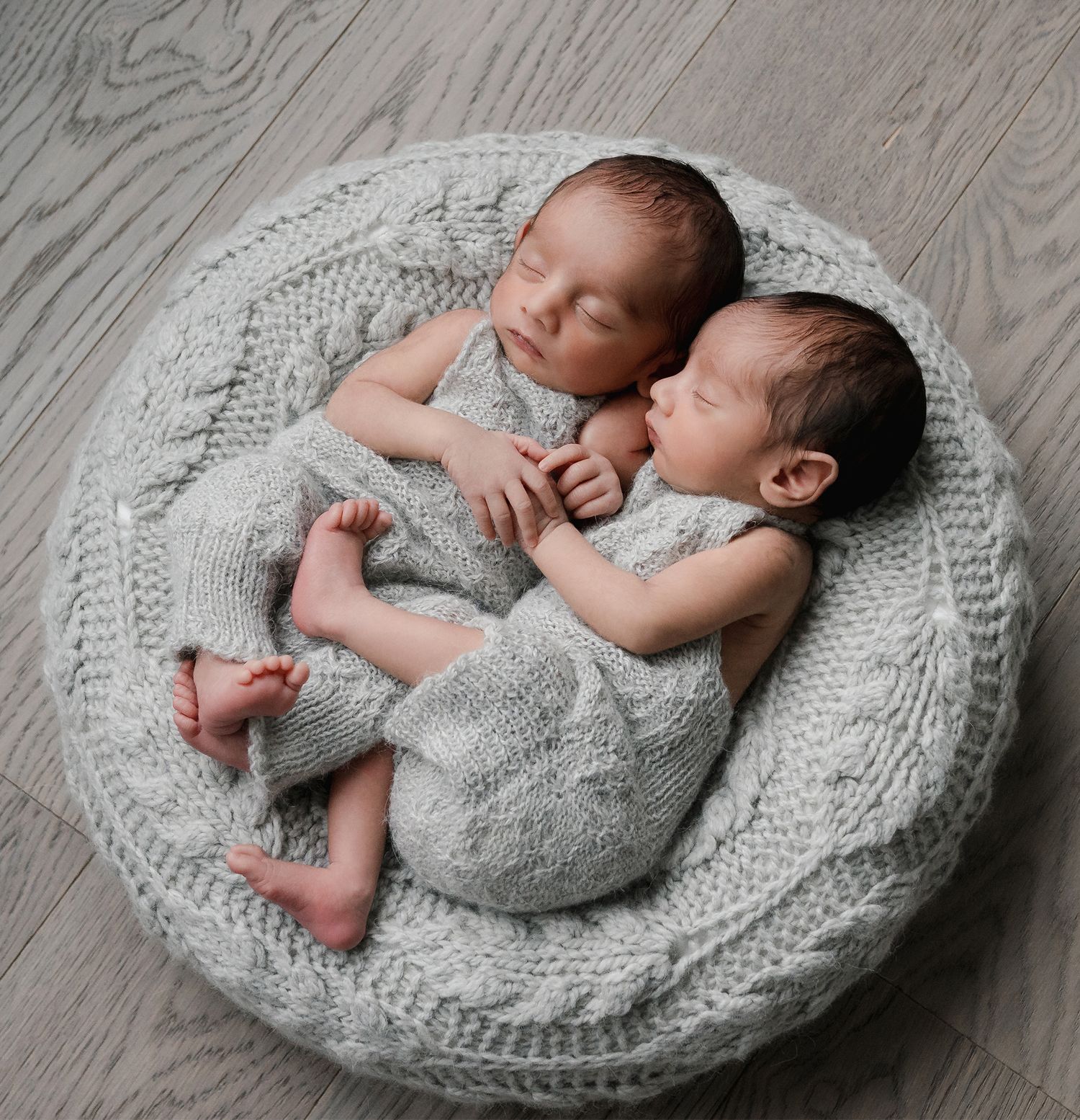 Twin newborn babies sleeping together in matching gray knit outfits in a round knit bowl prop.