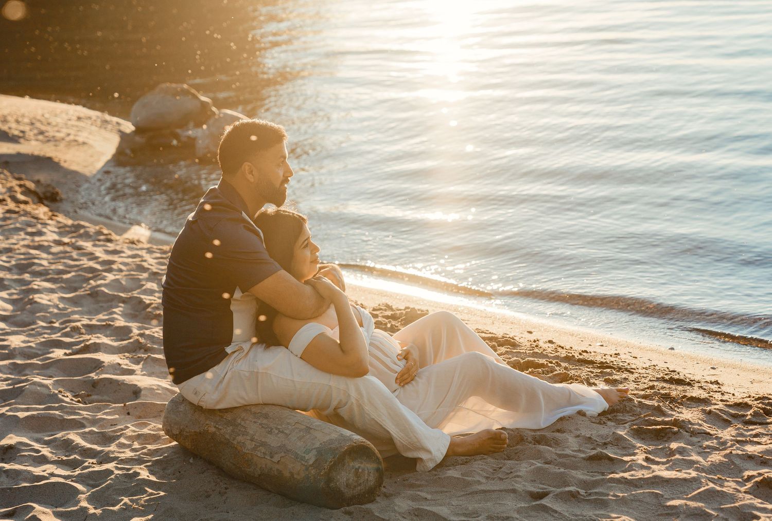 Man sitting on sandy beach at golden hour, gazing peacefully at calm sunlit water.