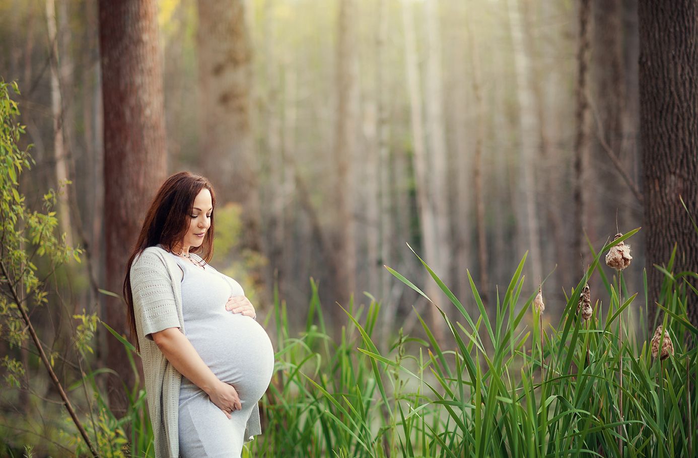 Pregnant woman in white dress cradling belly while leaning against tree in sunlit forest.