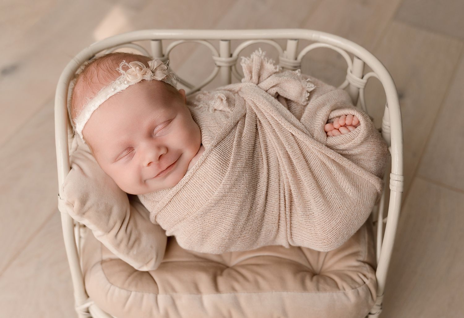 Smiling newborn baby in a cream knit wrap and floral headband sleeping peacefully in a vintage white metal bed.