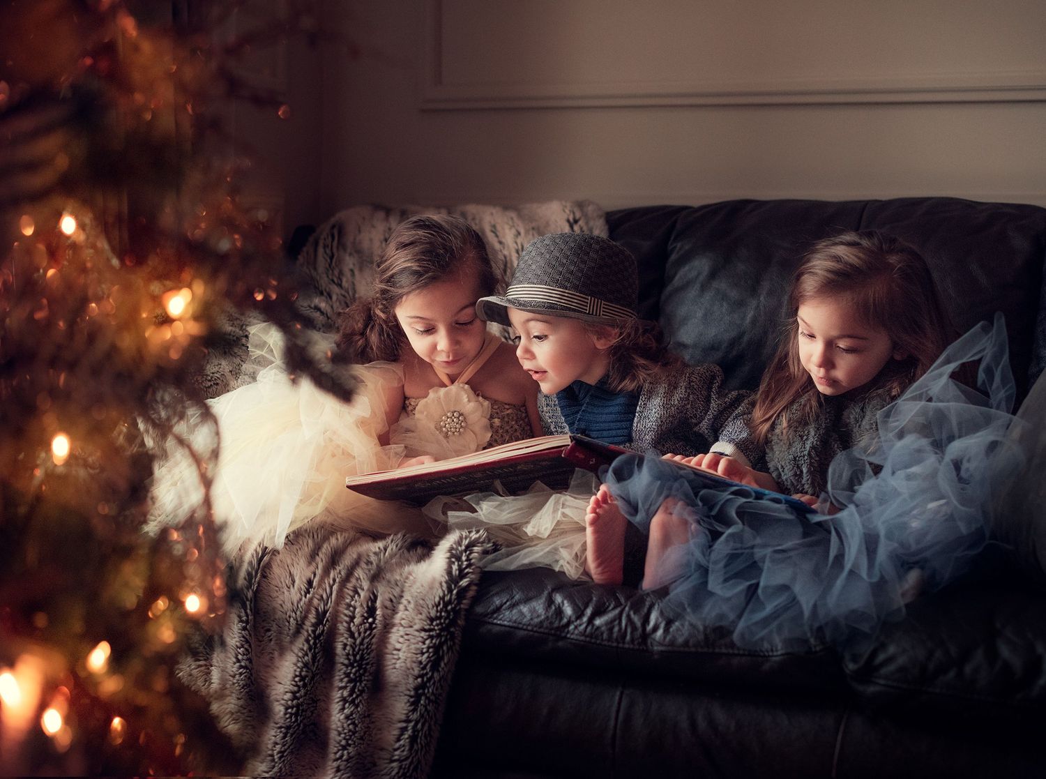 Three children reading a glowing book together on couch beside Christmas tree at night.