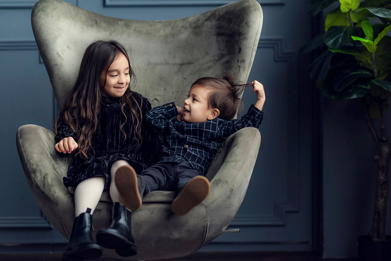 Two children sitting together in a large gray velvet egg chair, smiling and playing indoors.