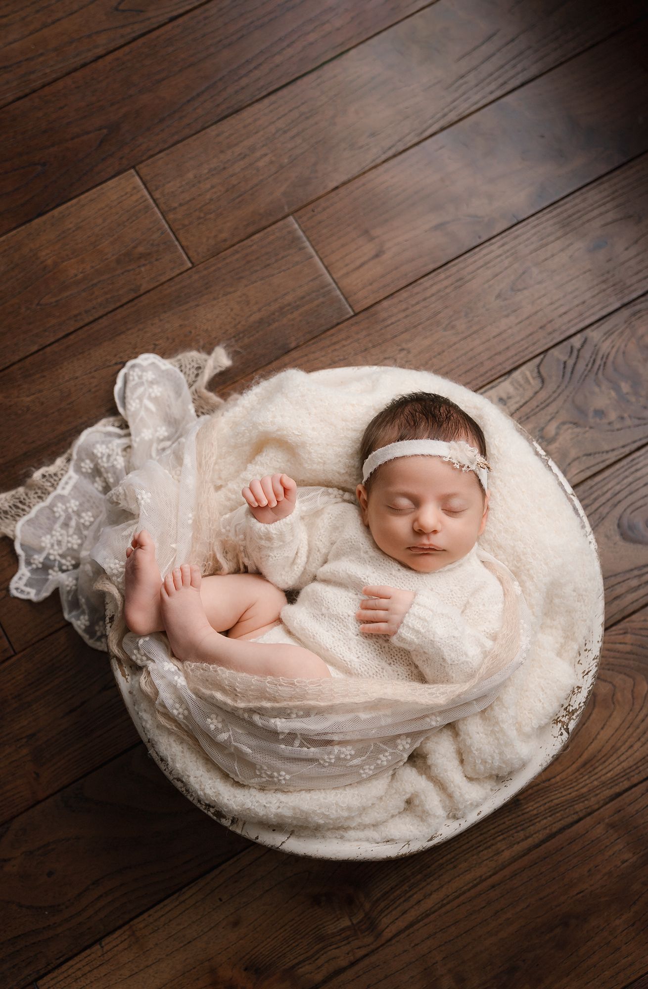 Newborn baby girl sleeping in a round bowl with cream lace wrap and floral headband.