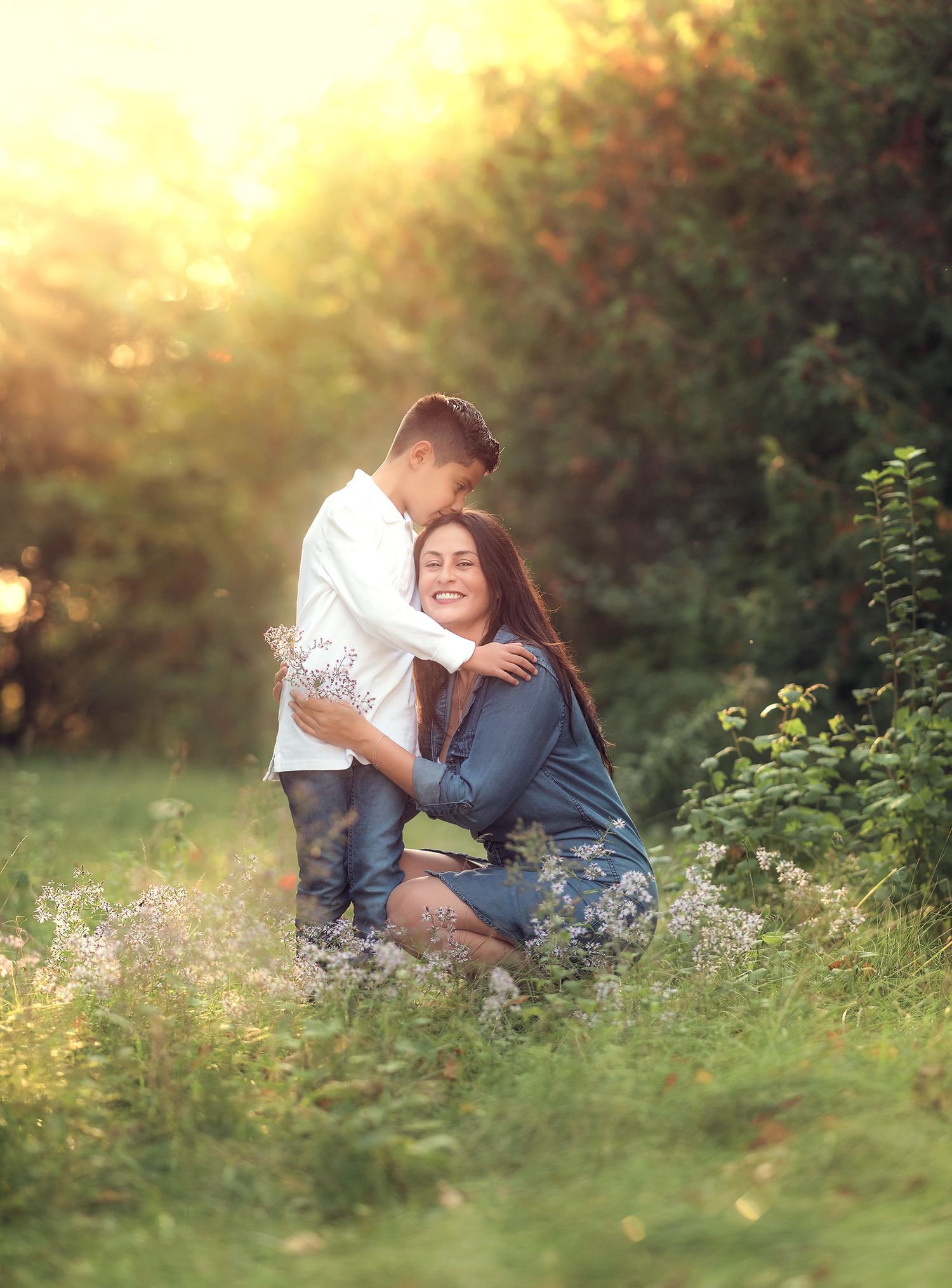 A smiling mother kneels in a sunlit meadow as her son embraces her from behind.