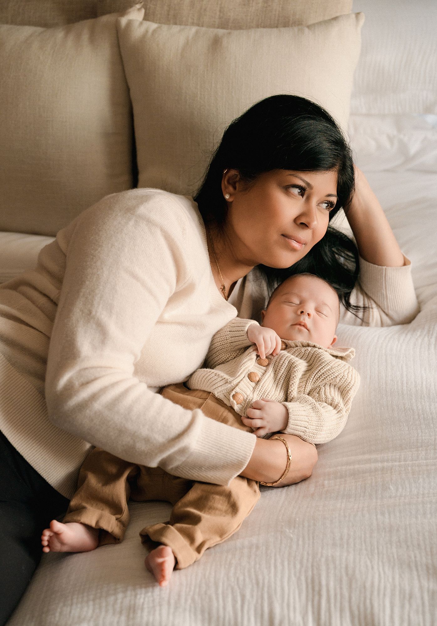 A woman in a cream sweater reclines on a bed, gazing away while cradling a sleeping newborn baby in a knit outfit.