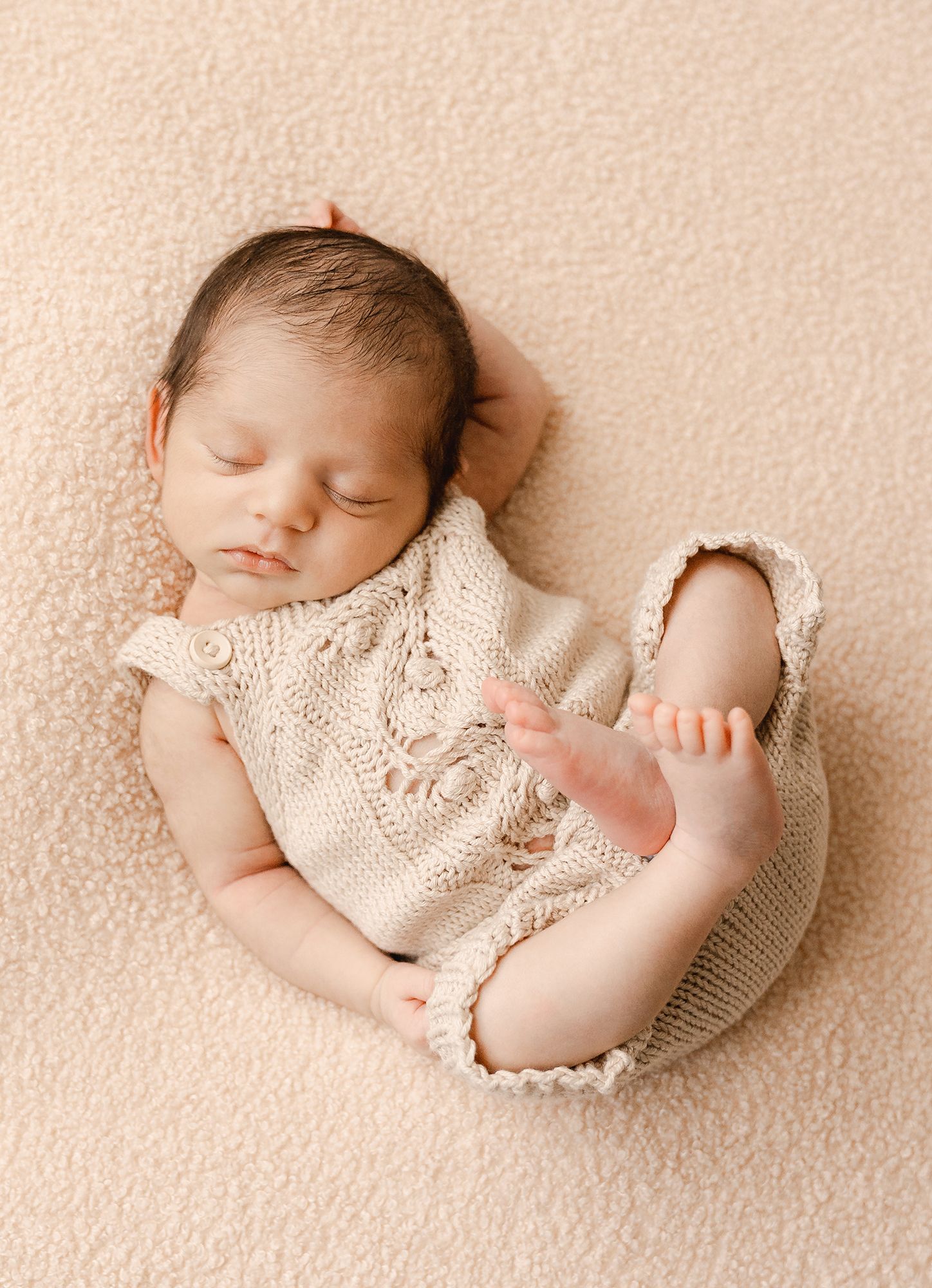 Sleeping newborn baby in a cream knit romper curled up on a soft beige blanket with tiny feet raised, a peaceful newborn photography pose.