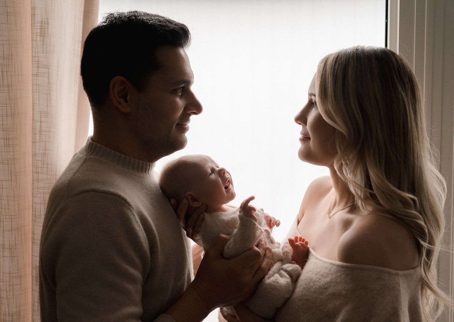 Silhouetted parents gaze lovingly at each other while the father cradles their sleeping newborn baby beside a bright window.