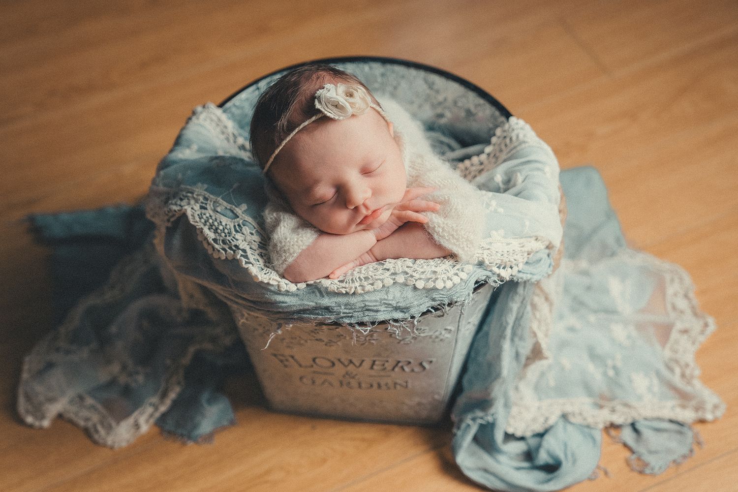 Peaceful newborn baby girl sleeping in lace-lined bucket with soft blue fabric decor.