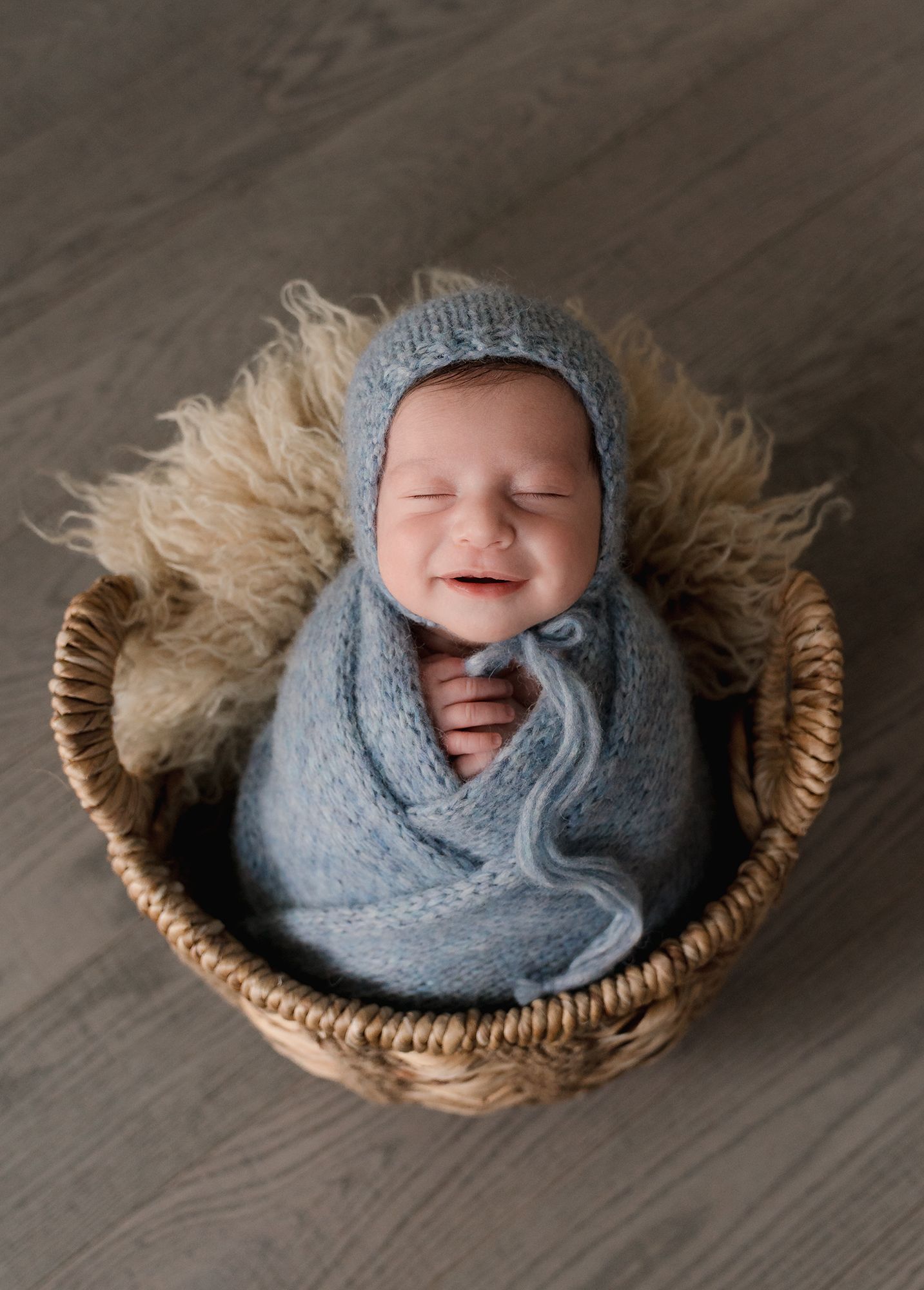 Smiling newborn baby wrapped in blue swaddle and knit bonnet sitting in a wicker basket.