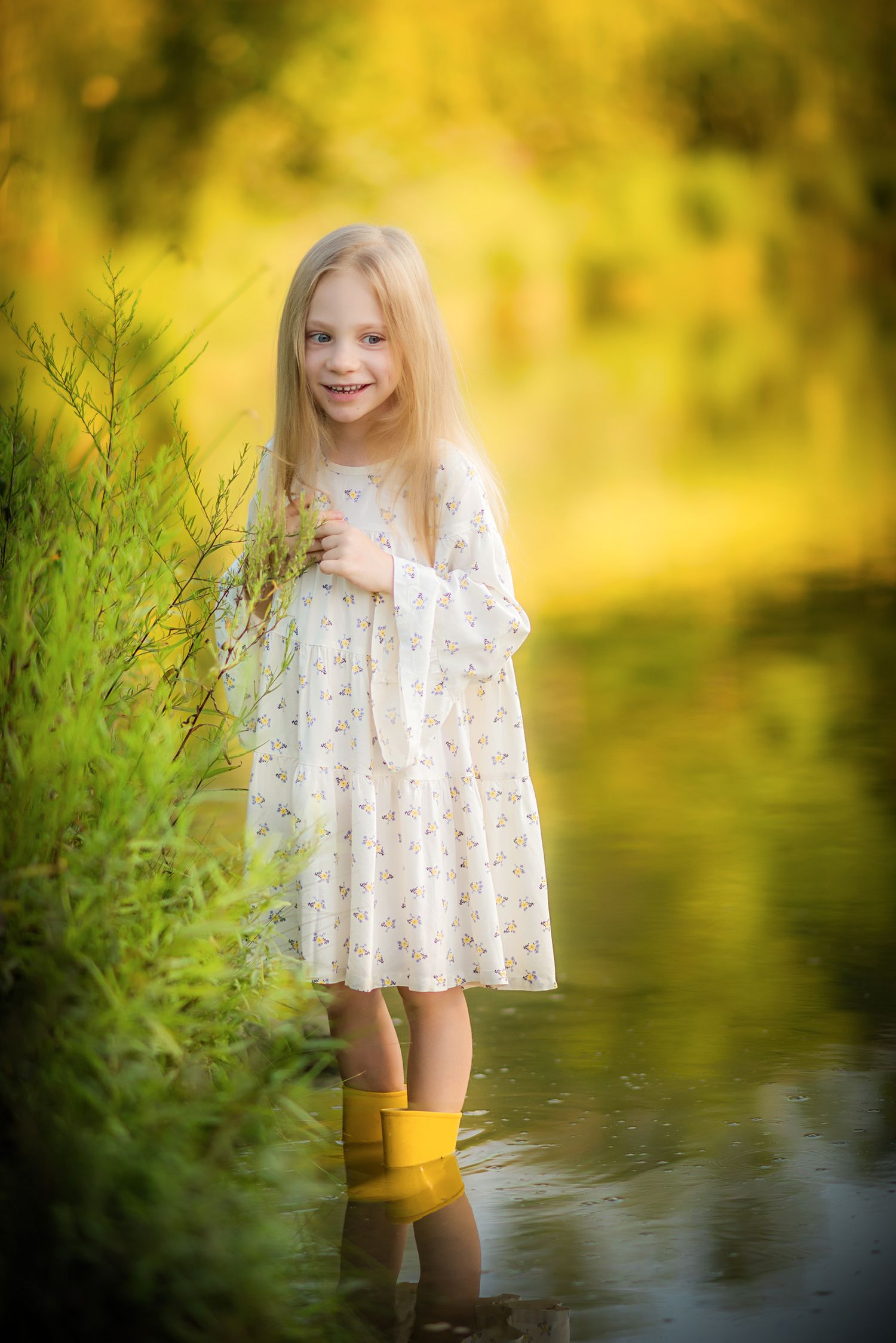A cheerful young blonde girl in a floral dress and yellow rubber boots stands in water outdoors.