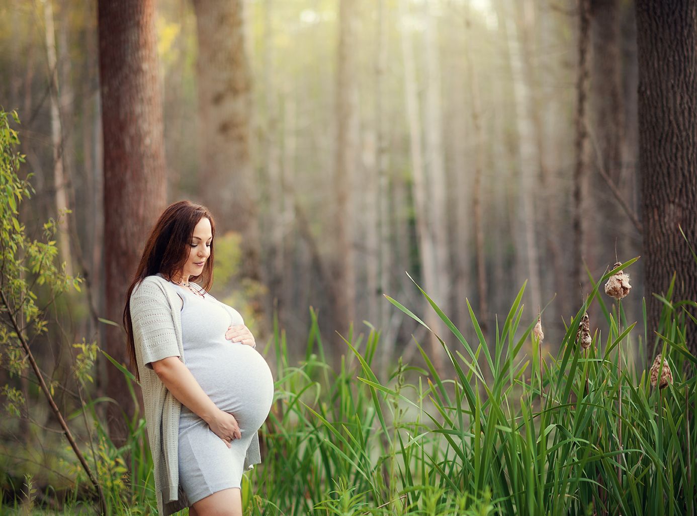Pregnant woman in white dress cradling baby bump in sunlit forest with tall trees and green grass.