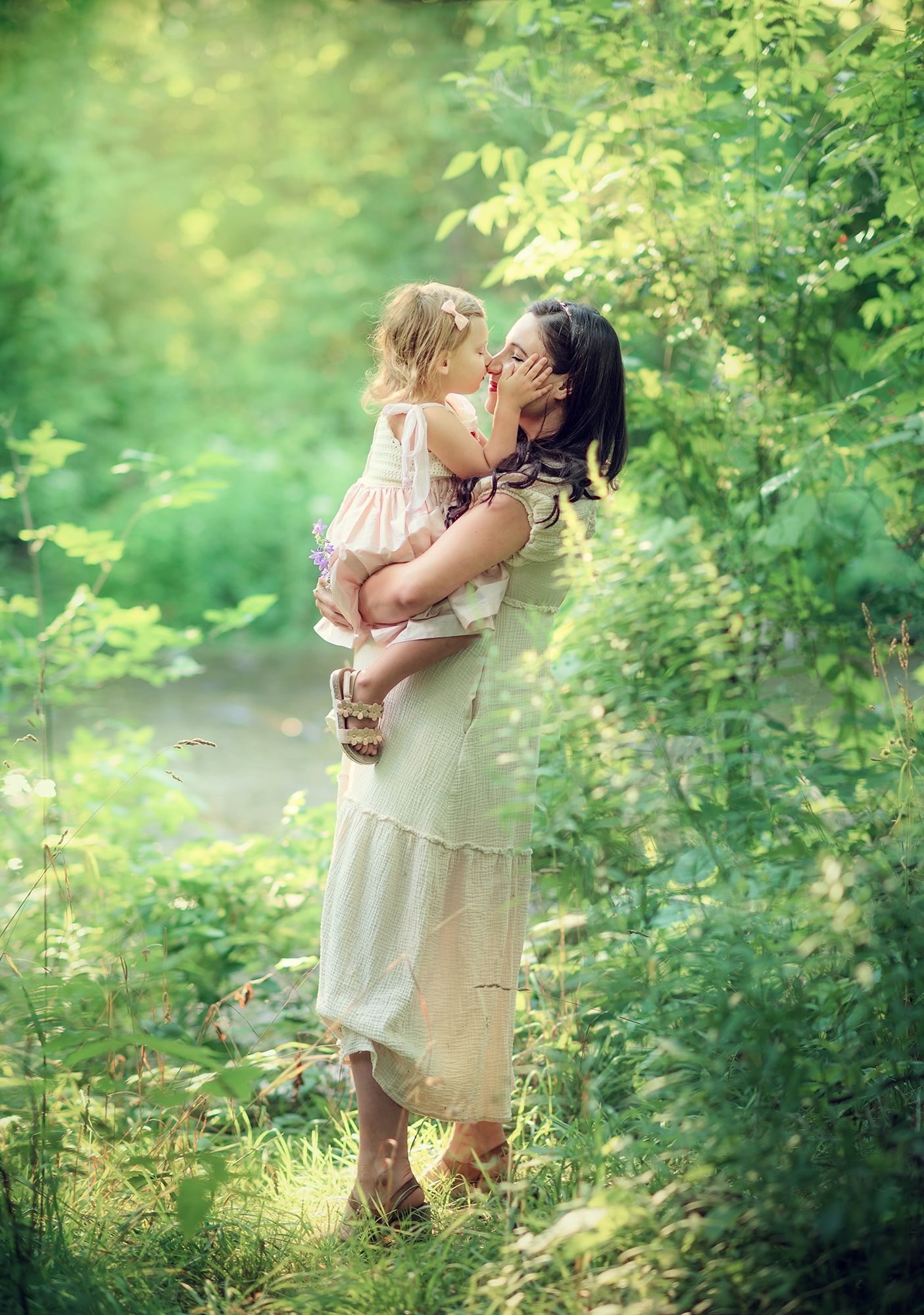 Mother in cream dress kissing toddler daughter while standing in lush green garden.