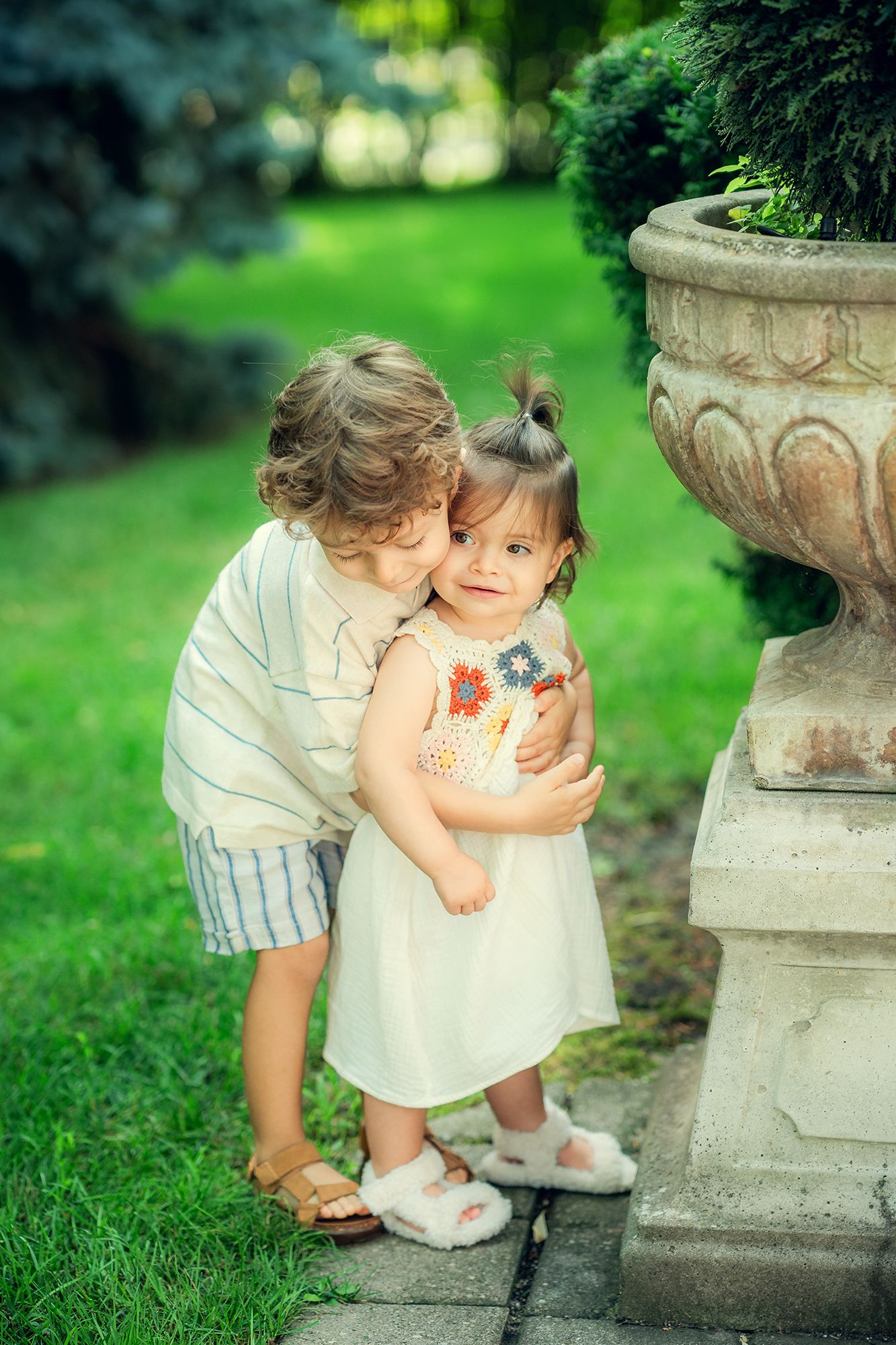 A curly-haired boy gently kisses a smiling toddler girl on the cheek in a lush garden.
