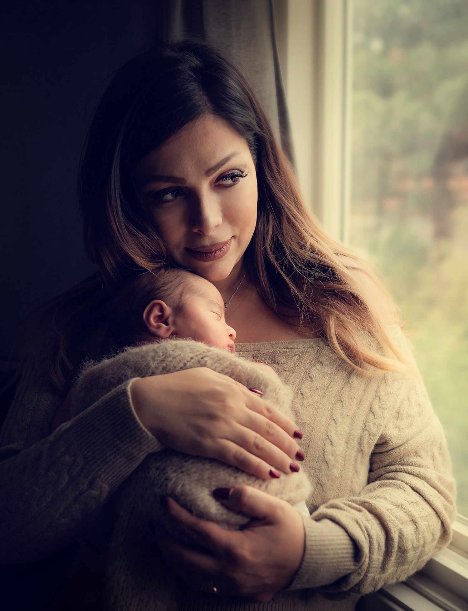 Mother holding her sleeping newborn baby wrapped in a knit blanket near a window.