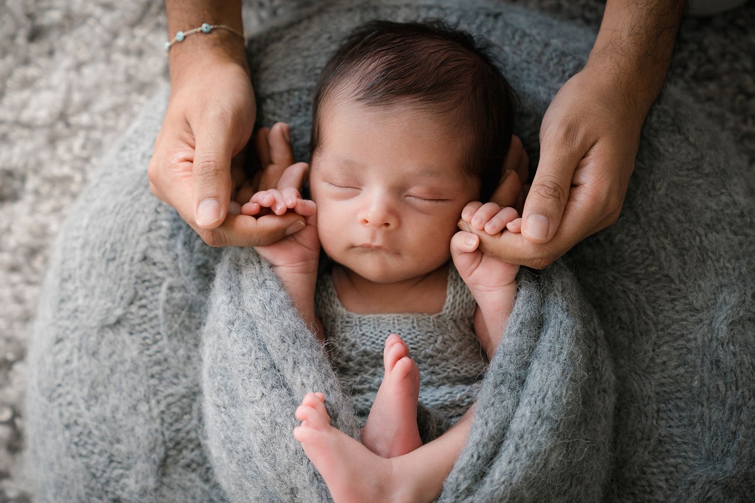 Parents gently holding sleeping newborn wrapped in gray knit swaddle from above.