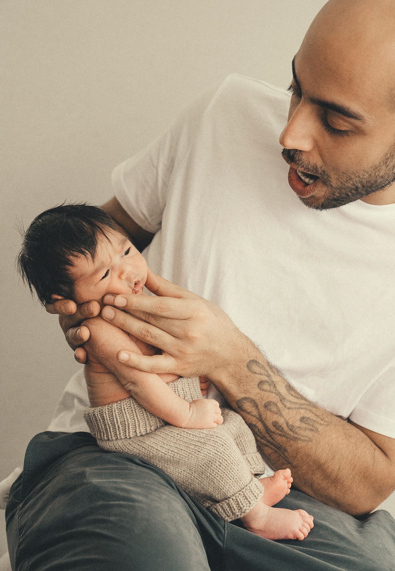 Tattooed father in white t-shirt holds sleeping newborn baby with dark hair on his lap.