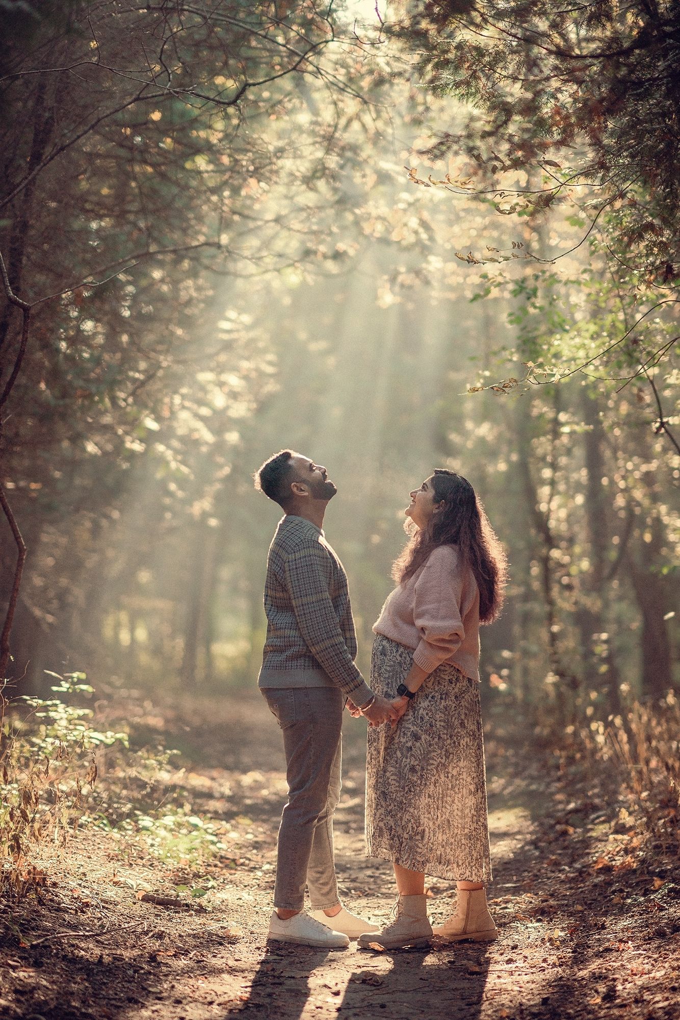 Couple holding hands gazing upward in a sunlit forest with golden rays streaming through trees.