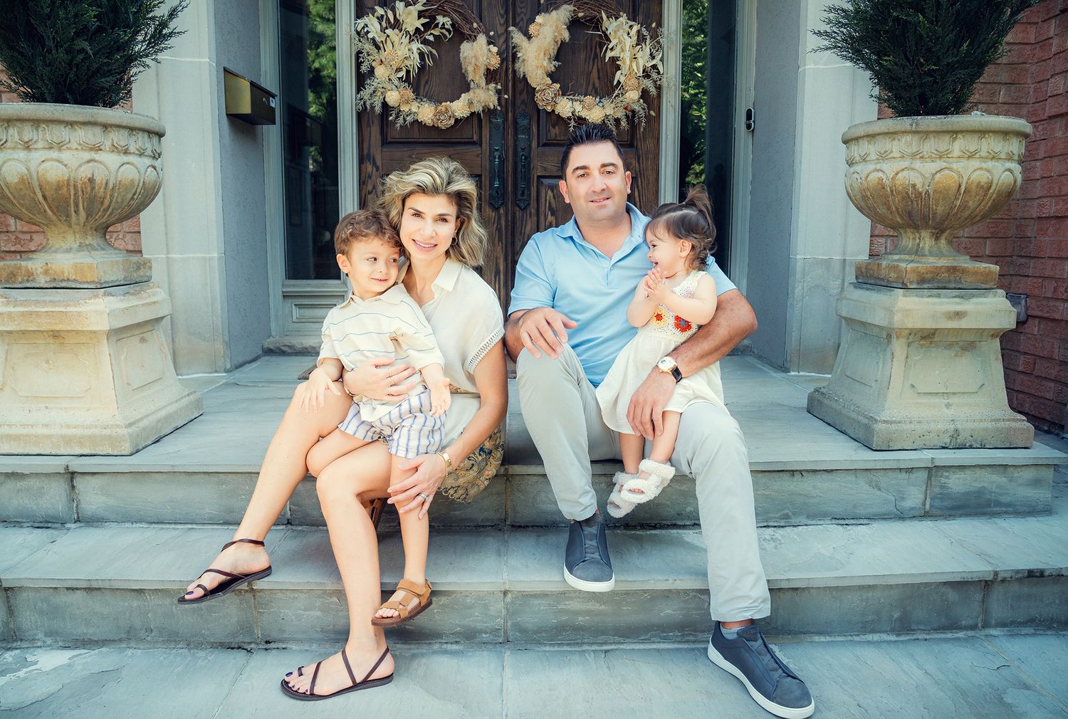 A couple with two young children sits on front steps of an elegant home with decorative urns.