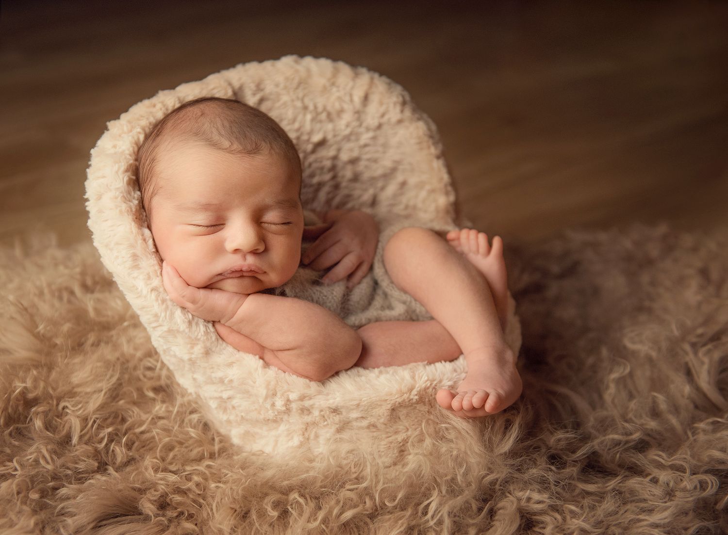 Peaceful newborn baby sleeping curled up in a fluffy cream bowl on a soft beige fur blanket.