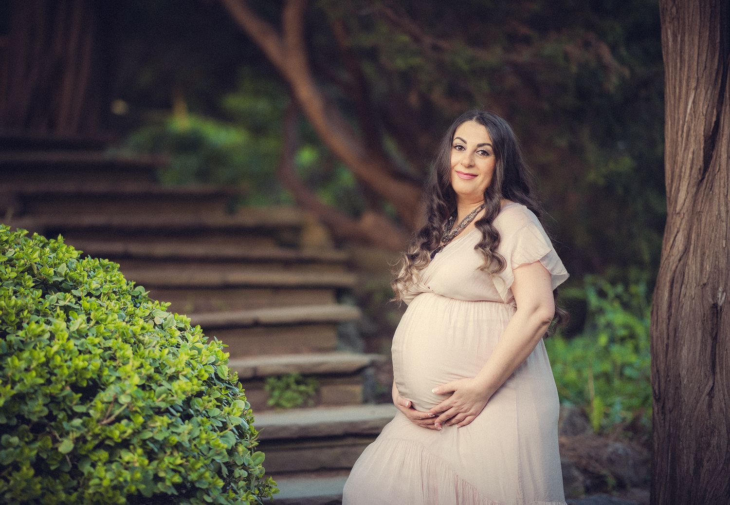 Pregnant woman in blush gown holding belly near stone steps and green bushes.