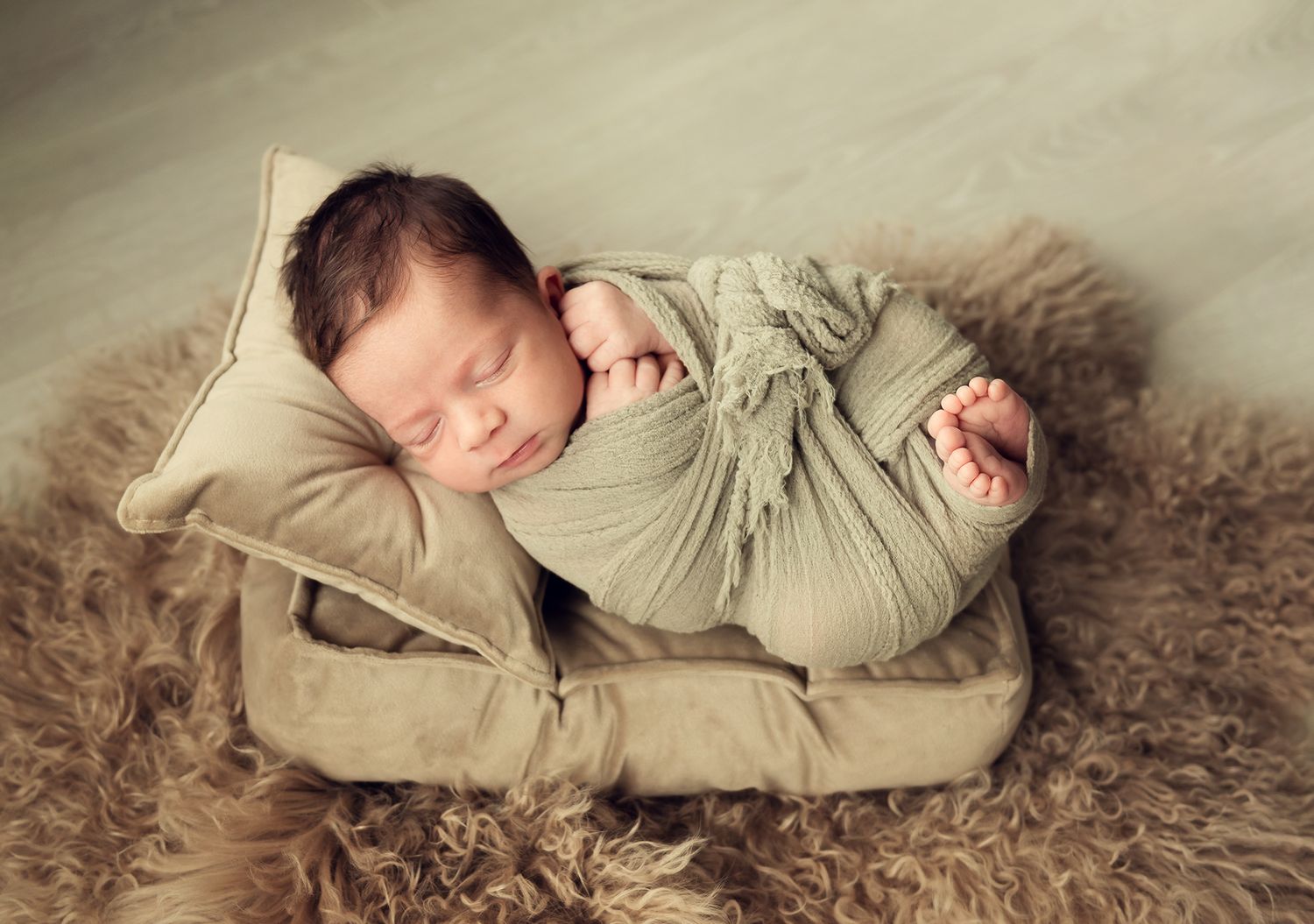 Sleeping newborn swaddled in a sage green wrap resting on a tiny pillow bed on fur.