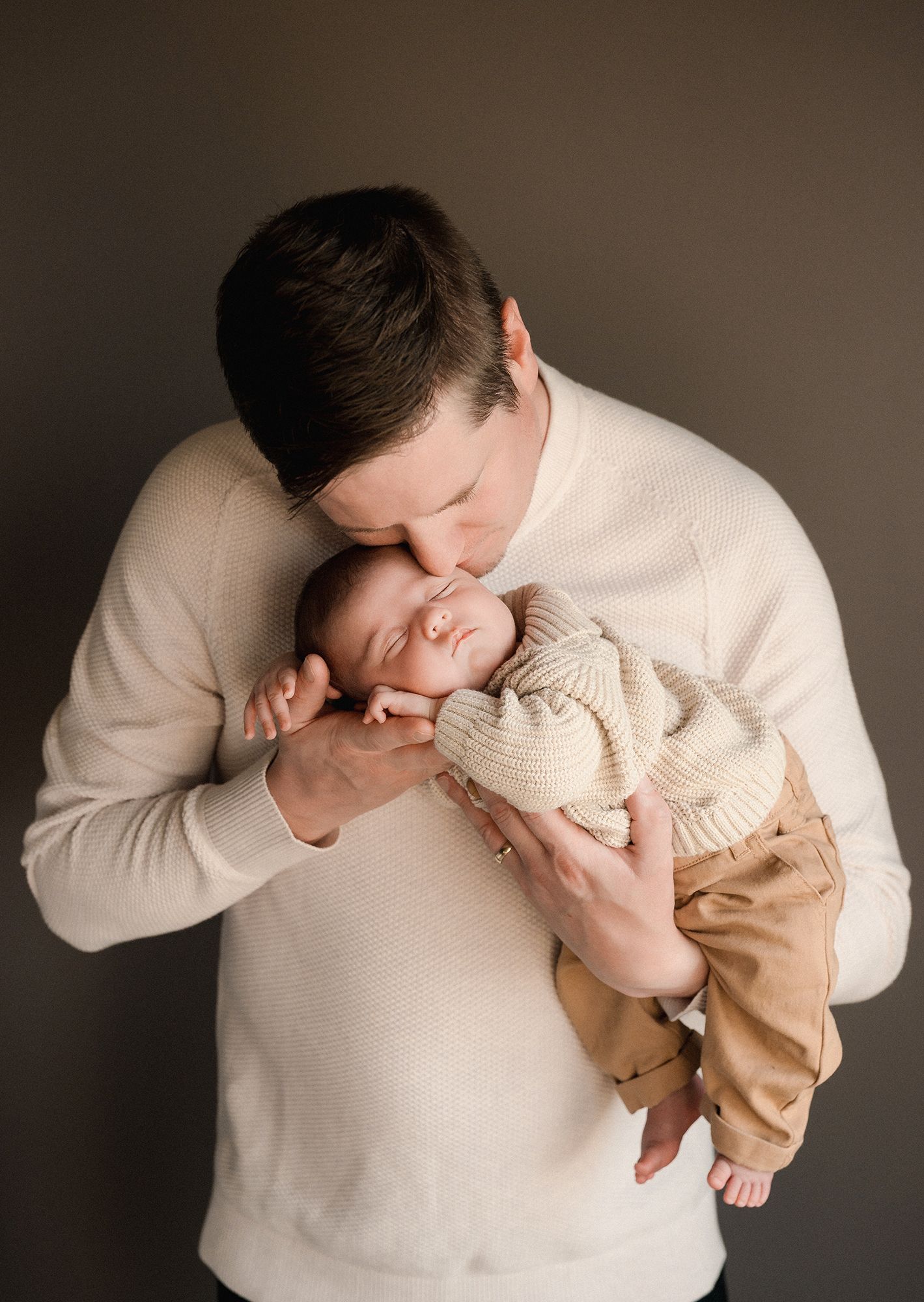 A father in a cream sweater gently kisses the forehead of his sleeping newborn baby dressed in a knit top and tan pants.