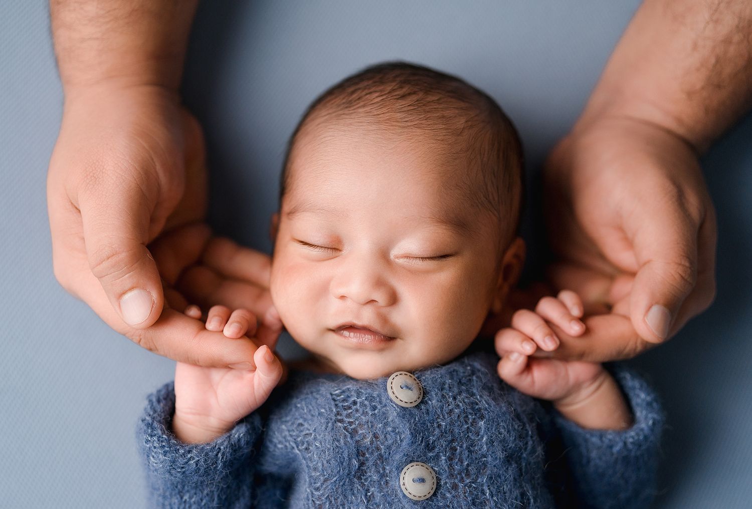A smiling newborn baby in a blue knit cardigan rests gently in his fathers cupped hands.