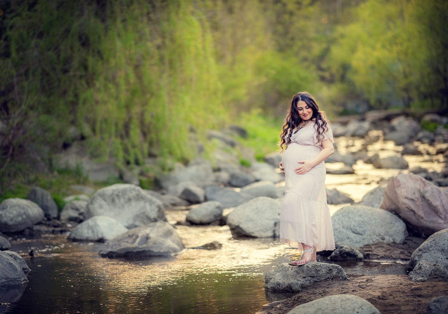 Pregnant woman in white dress standing on rocks beside a serene forest river.